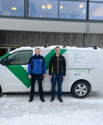 Dette er en bil med to personer foran. Bilen er en varebil, og har 4 hjul og grønn logo